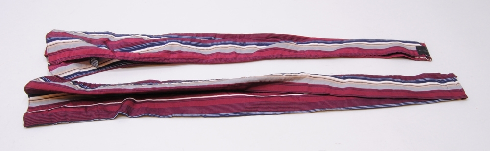 Scarf, tvådelad med kardborrknäppning i nacken. Scarfen är tillverkad av randigt polyestertyg i vinrött, grått, mörkblått, beige och vitt. Ränderna har olika tjocklek. Till scarfen hör ett scarfsmycke, se Jvm 21059.