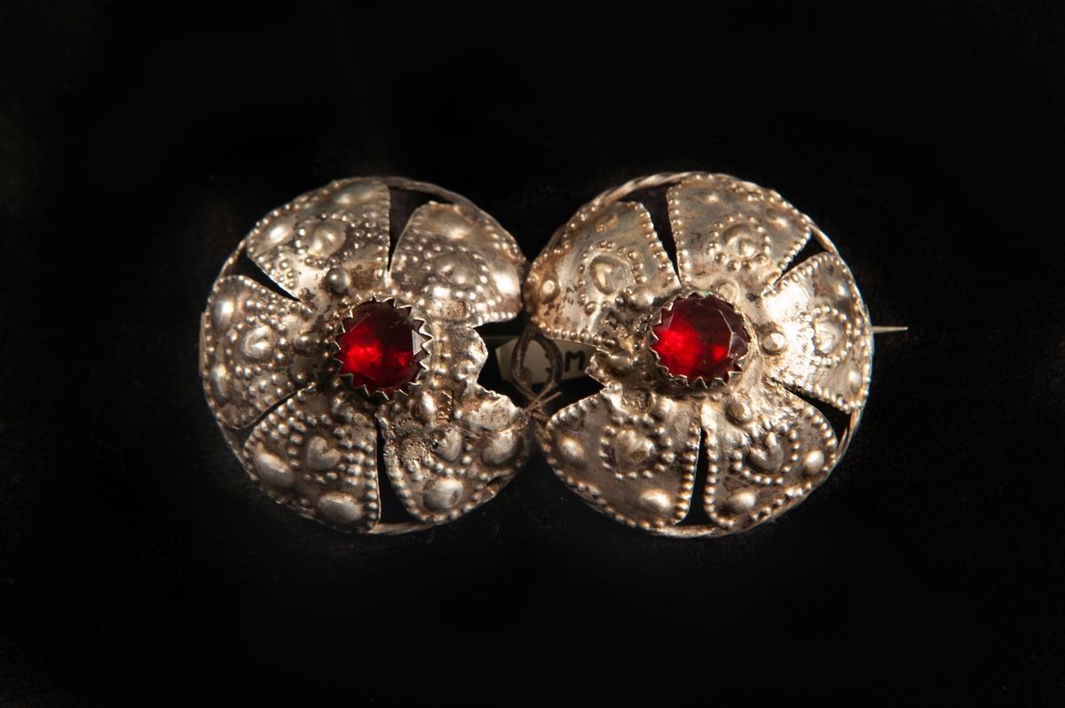 Två hoptvinnade maljor av ciselerat silver med dekor av hjärtan m.m. och med två infattade stenar av rött glas. Maljorna har gjorts om till en brosch.