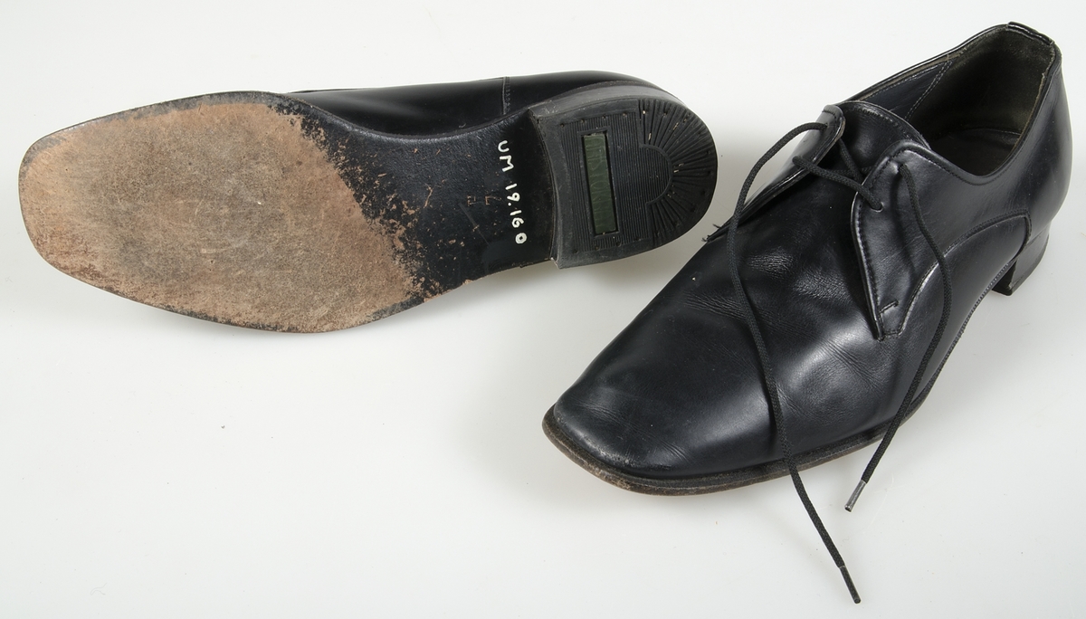 Ett par snörskor av svart maskinsytt läder och svarta snören. Fyra snörhål på varje sko. Sulor av svart gummi. Räfflade klackar. Kraftigt avsmalnande mot tårna. Märkt otydligt på insidans häl och utanpå klacken: ARISTOKRAT. Samhörande med nr. UM 19115-19188.