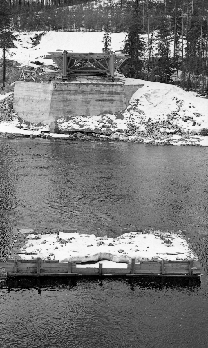Fra bygginga av Øvre Flisbru i 1961. Fotografiet er altså tatt ved elva Flisa i Åsnes i Solør, antakelig like nedenfor Vålbekken, der Linnavegen skulle krysse vassdraget og forene seg med Gravbergsvegen på vestsida av vassdraget. Fotografiet er tatt på tvers av elveløpet. I forgrunnen ser vi et forskalet, uferdig fundament, og på motsatt side av elva var det et ferdigstøpt brukar med ei påbegynt treforskaling - antakelig til sjølve vegbrua - på. Prosjektet interesserte antakelig fløtingsingeniøren som har tatt dette fotografiet fordi den bærekonstruksjonen som etter hvert skulle reises på det støpte fundamentet sentralt i elveløpet kunne bli et hinder for framdriften av fløtingstømmeret i Flisa. Stokkene hadde lett for å hekte seg fast i slike brukar.