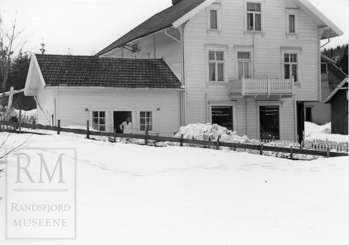 Vinterlandskap. Ouliebutikken, Ouliegården, nordveggen: Mjølkemottak til venstre. Arne Oulie står i døra..