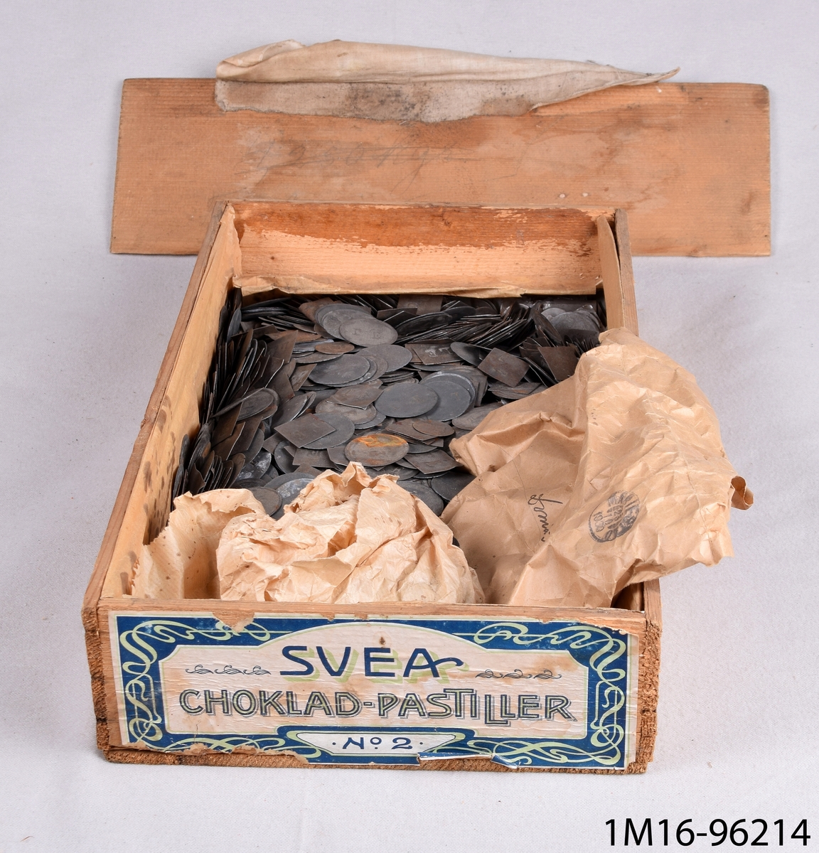 Plåtbrickor, runda, fyrkantiga, som byttes mot pengar. Ligger i en låda av trä med etikett med texten Svea Chokladpastiller.