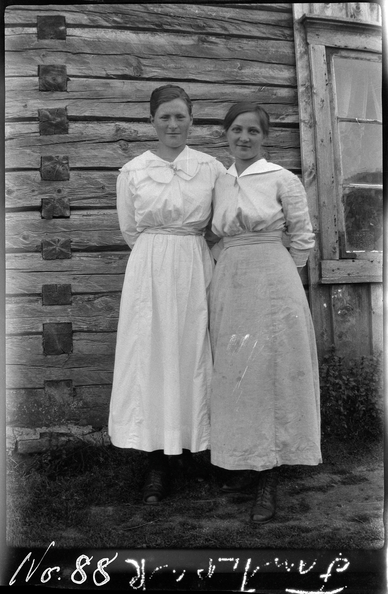 Portrett av to kvinner stående foran en laftet husvegg.
