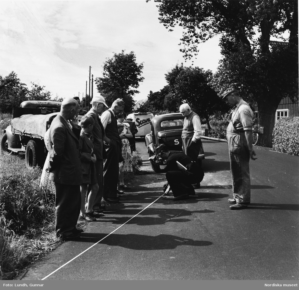 Trafikolycka i Skåne år 1950. En polis mäter med måttband på vägen medans en folksamling tittar på.  Bilar och en lastbil står vid vägkanten.