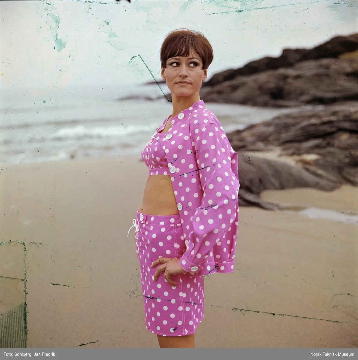 Kvinnelig modell viser bademote, hun har shorts, bikinioverdel og jakke, alt i rosa med hvite prikker. Hun er fotografert på en strand.