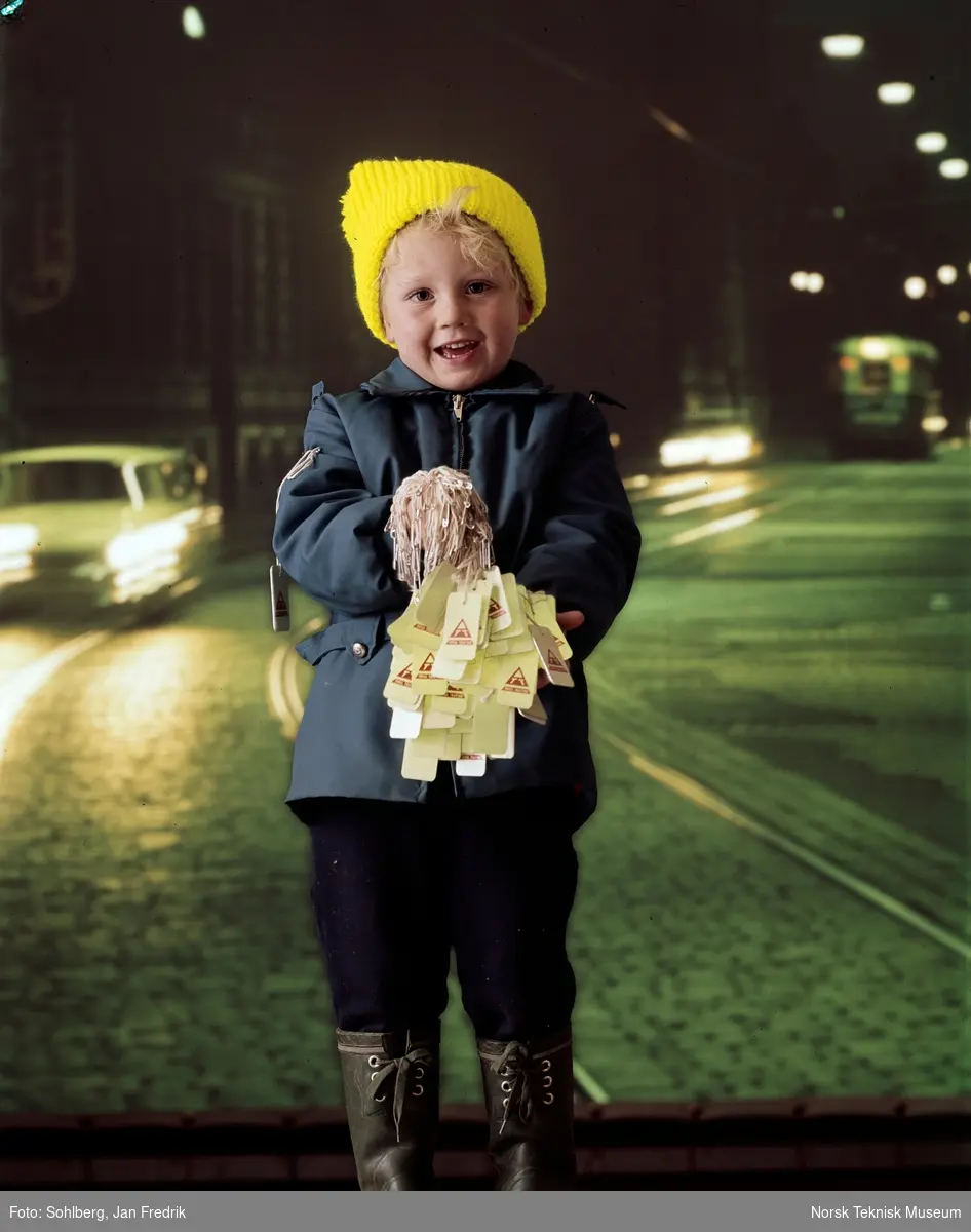 En liten gutt med gul toppelue og blå anorakk holder fram mange reflekser med reklame for Trygg trafikk. Fotografert i studio, bakgrunnen forestiller en mørk gate.