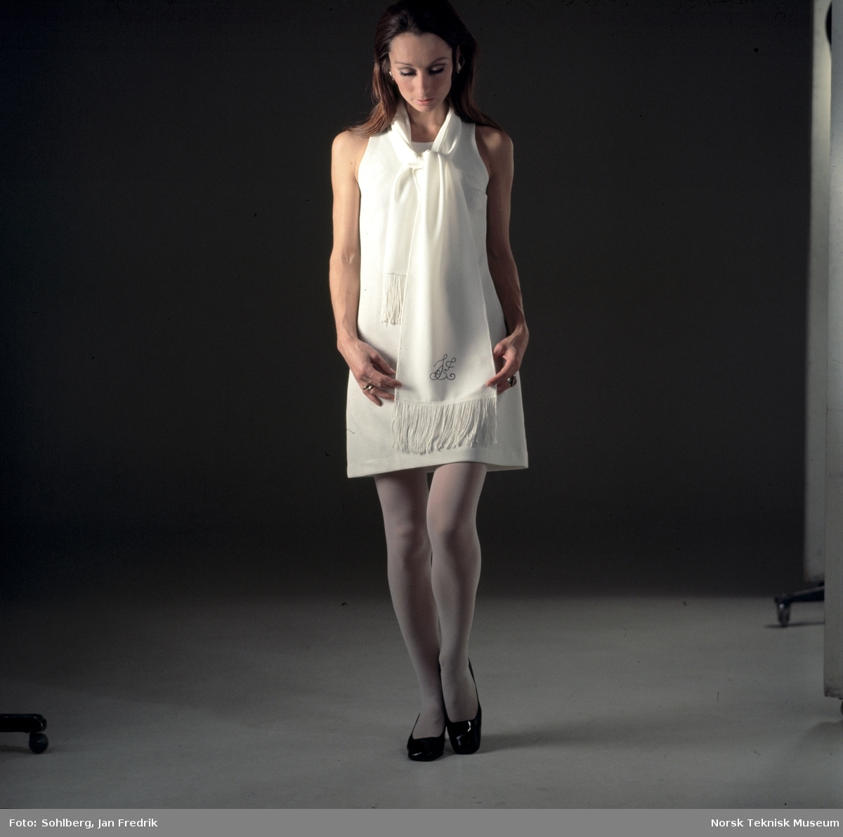 Tidlig norsk motefotografi. Kvinnelig modell med kort, hvit kjole og skjerf med monogram.