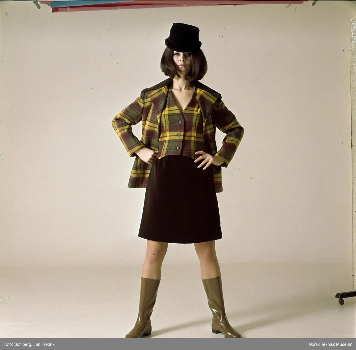 Tidlig norsk motefotografi. Kvinnelig modell med rutete jakke og vest i gult,  brunt og rødt og et mørkebrunt skjørt. Tlbehøret er en tøff hatt og lysebrune gummistøvler.