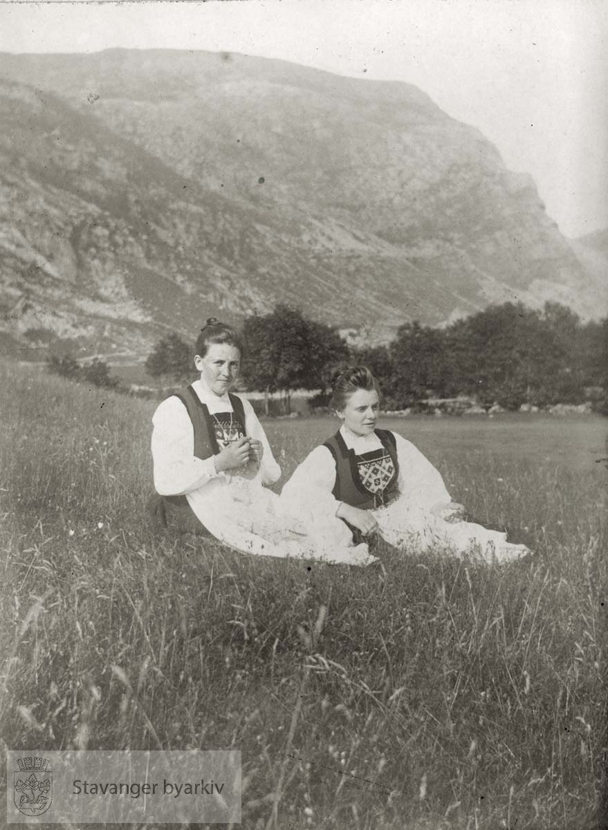 To kvinner i bunad sitter i en eng. Fjell i bakgrunnen.