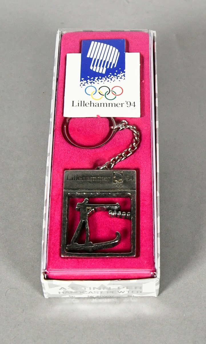 Nøkkelring med rektangulært anheng med piktogram for skiskyting. Nøkkelringen ligger i original emballasje på rosa underlag.