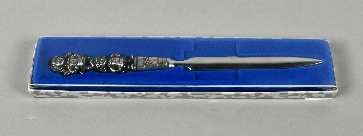 Brevkniv av tinn med maskotene Kristin og Håkon ytterst på skaftet. Brevkniven ligger i origianl emballasje på blå bunn.