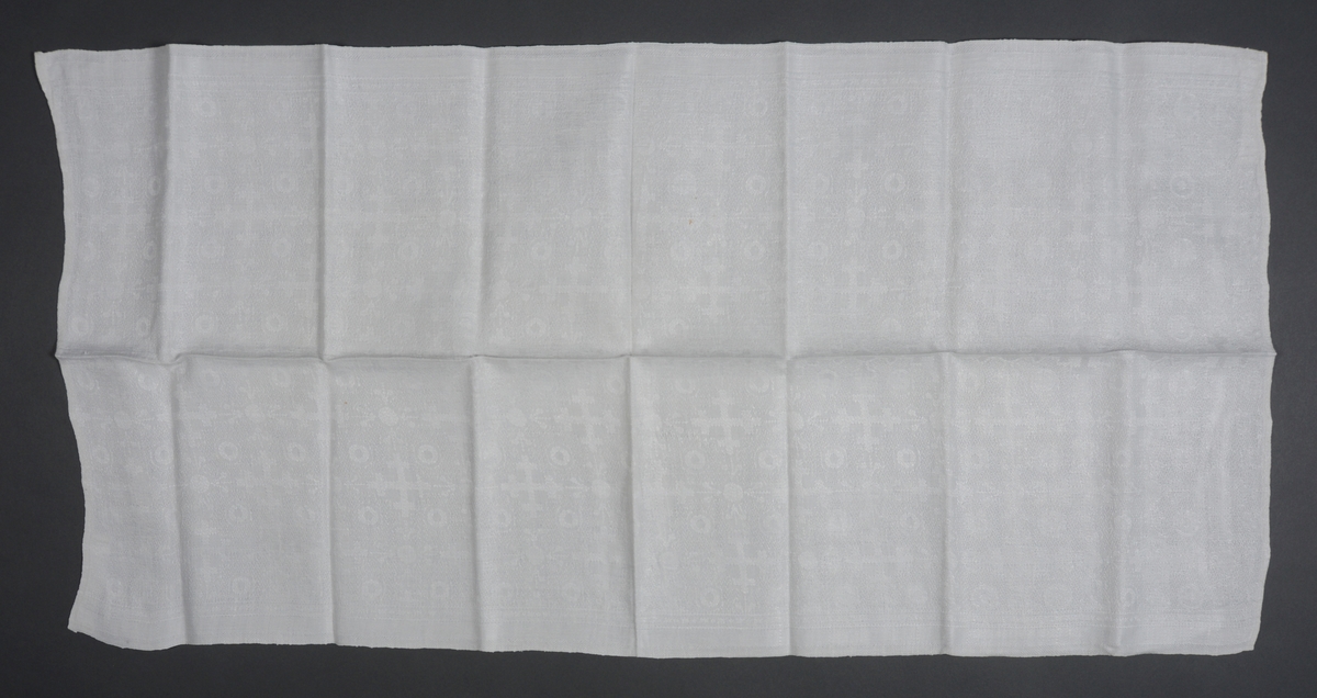 Rektangulær damaskvevd håndduk. Kortsidene på stoffet er kantet inn med maskin. Påsydd hempe av bomullsbånd på begge kortsidene.