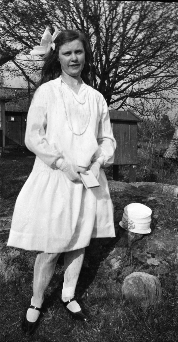 Flicka som står utomhus i konfirmationskläder, långt pärlhalsband och stor rosett i håret. Både klänningen med lågt skuret liv och hatten på stenen bredvid henne är av 1930-talssnitt.