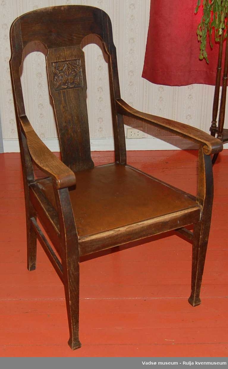 Bred stol med lærtrukket sete. Ryggstø er buet i topp. Åpen rygg med vertikalt element i midten. På dette er et kvadratisk felt med utskjært bladdekor, ca. 15 x 15 cm.