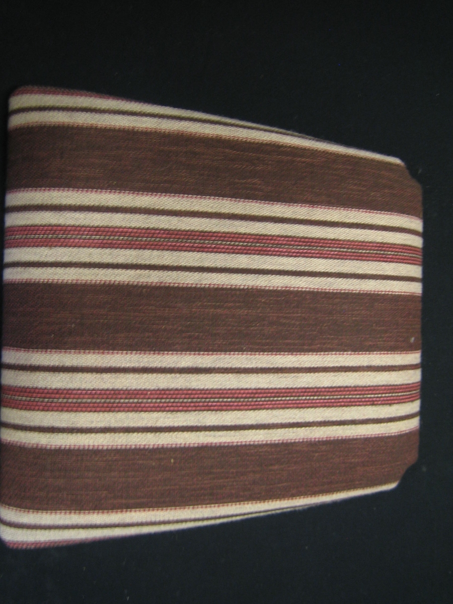 Möbelöverdrag, möbeltyg monterat på stolsdyna. Ett stadigt möbeltyg i tuskaft Tyget är randigt med breda ränder i brunt och smalare i grått, brunt och rosa. Varpen är av rött bomullsgarn. Inslaget brunt, grått och rött ullgarn och rosa lingarn.

Möbeltyget är monterat med häftklammer på en stolsdyna av skivmaterial av trä med stoppning. Dynan är lite smalare i bakkant och där är hörnen urtagna. Dynan är monterad av TRÄ-TINA i Lerdala. På undersidan sitter en vit klisterlapp med texten: möbeltyg "Åker". Kvalitet Inslag - ylle-lin, Varp - bomull, Bredd 131 cm, Pris 380:-
SKARABORGS LÄNS HEMSLÖJDSFÖRENING och en lapp med texten: "TRÄ -TINA, Storekvarn Pl 397, 540 17 LERDALA, 0511-804 64
Det finns också ett vävprov märkt RSLH-0681-1