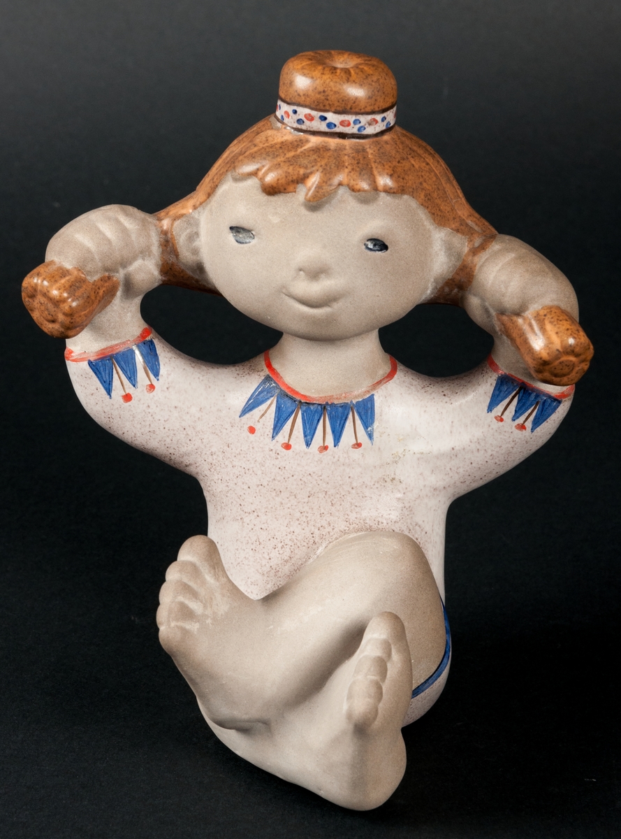 Figurin i flintgods föreställande flicka sittande med benen i kors. Rödhårig med knut på huvudet och flätor som hon håller i med varsin hand.