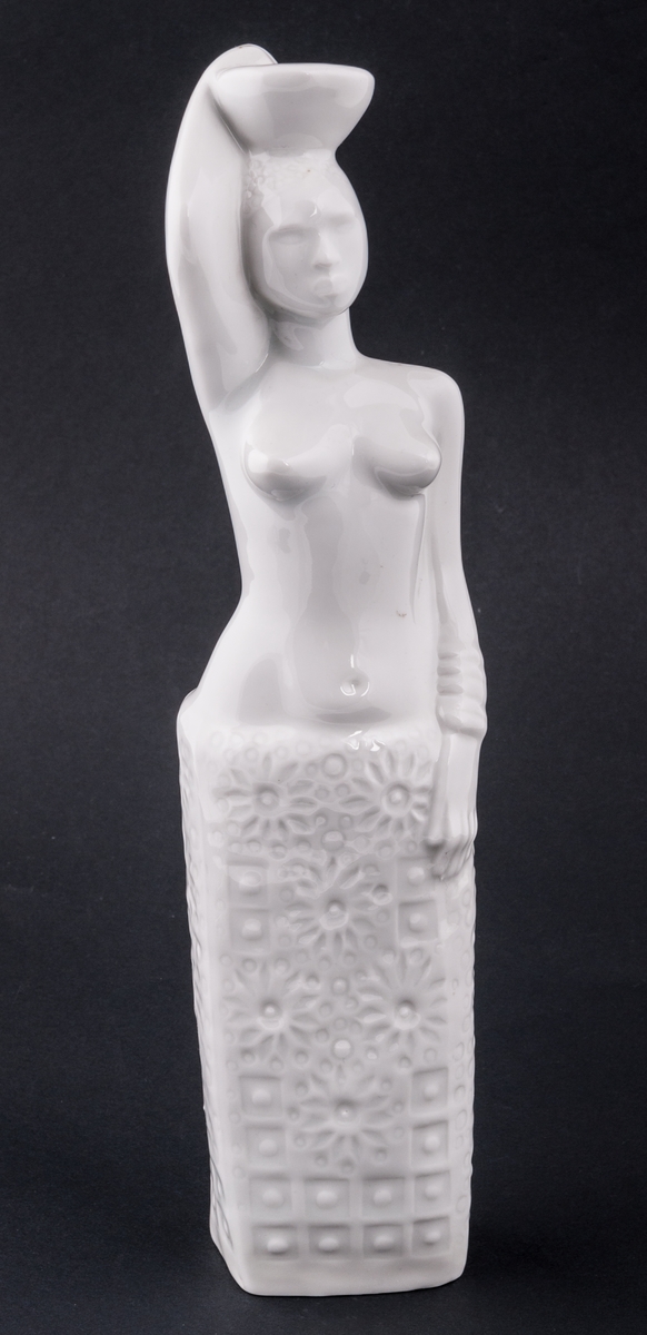 Figurin, föreställande en kvinna, bärandes kärl på huvudet, bar överkropp. Vit glasyr. Saknar stämplar och signatur. Okänd formgivare. Oklar tillverkare.