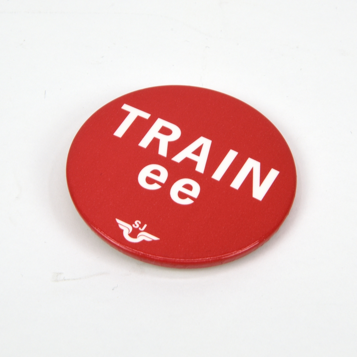 Rund pin med texten "TRAINee" i vitt mot röd botten. Under texten finns SJ:s logotyp. Baksidan består av vit plast med en infälld säkerhetsnål.