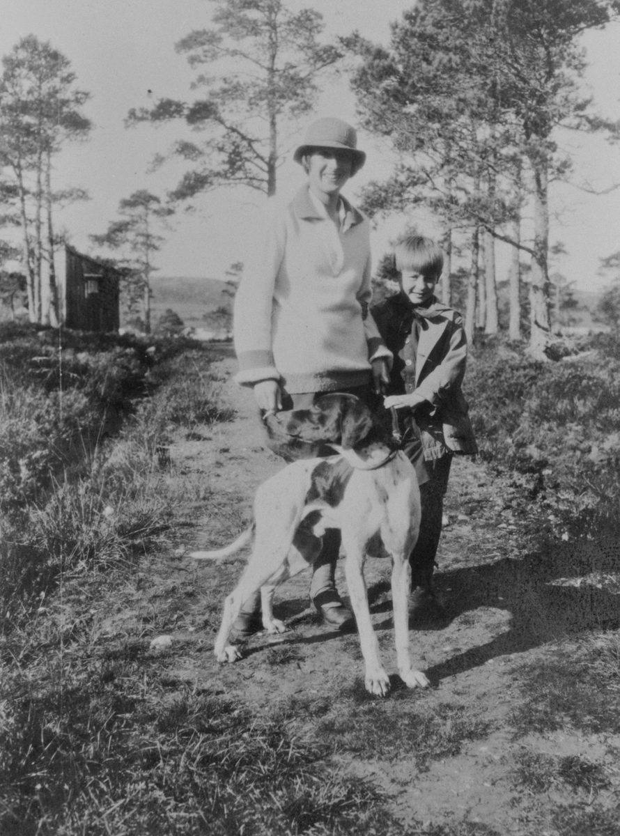 Lizzie Gran g. Juul og Stig Juul (hennes stesønn) på fottur med en hund, et uthus i bakgrunnen.