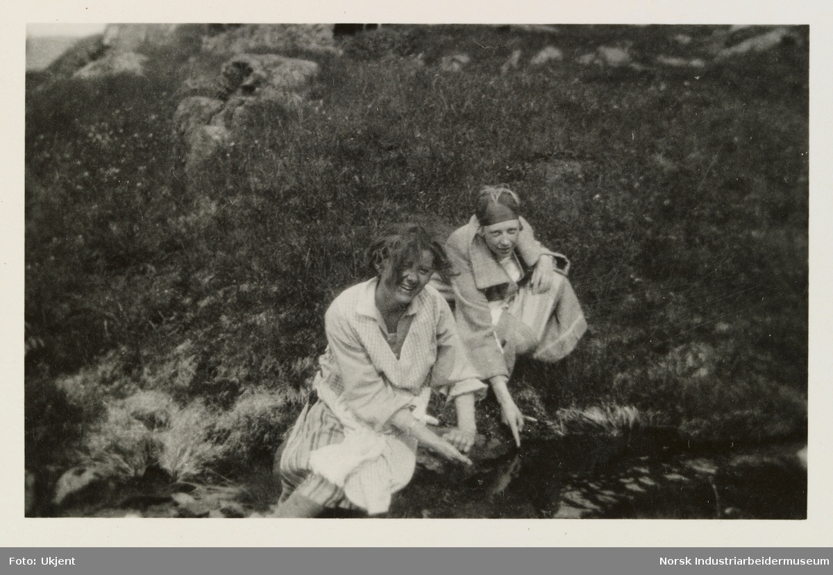 Kvinne og jente sitter på gress ved vannkant og peker ned mot vannet