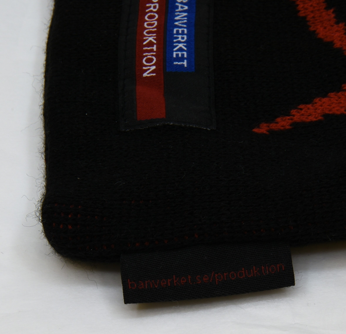 Maskinstickad mössa i svart med rött mönster på framsidan, samt en svart lapp fastsydd under mönstret. Vid mössans söm sitter ytterligare en svart lapp. Inuti mössan sitter ett tillverkarmärke.