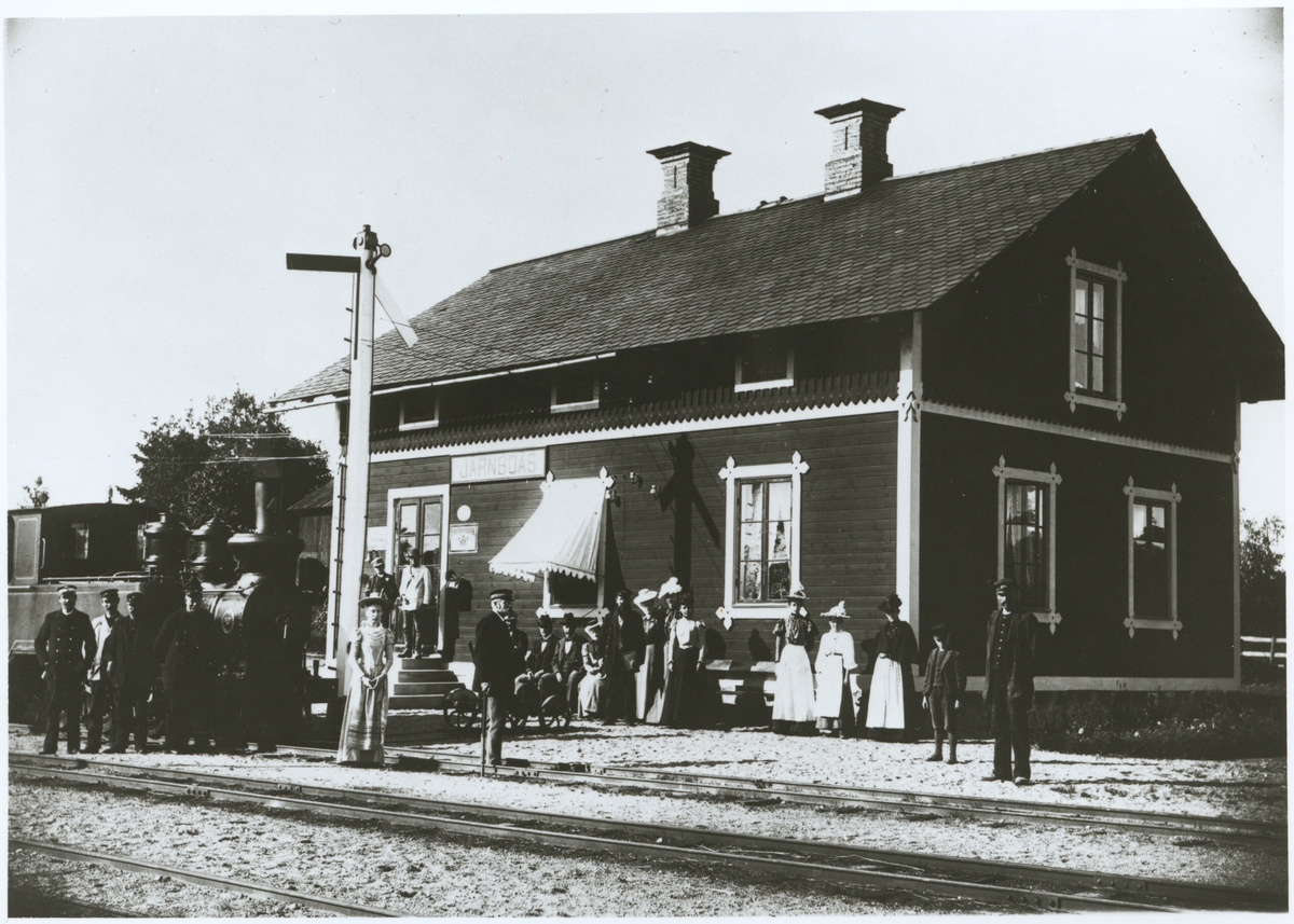 Järnvägsstationen i Järnboås med kvinnor och män som poserar utanför. Bredsjö - Degerfors Järnväg, BDJ lok 4 till vänster.