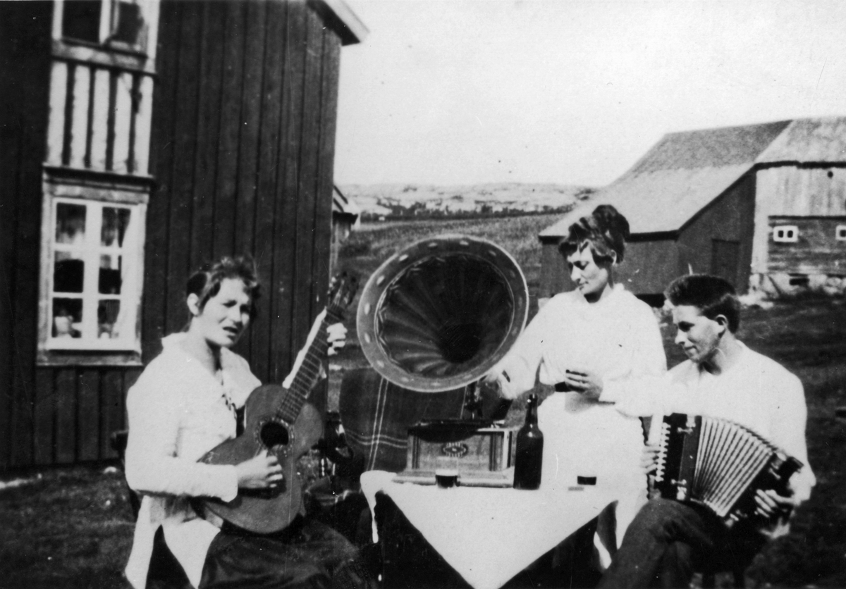 Musikkstund på gårdstun med gitar og torader. På bordet i midten står en sveivegramafon.
Bildet anskaffet i forbindelse med utstillingen 
"Spæll åt mæ", 1982.