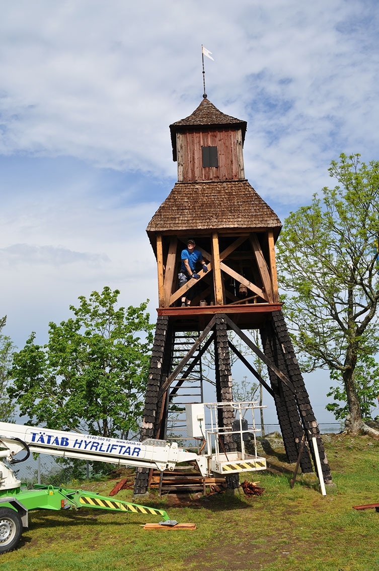 Klockstapeln vid Friluftsmuseet på Grännaberget. Panelen på klockhusets nedre del mot öster är nedriven. I öppningen syns Jari Telin. En skylift står framför klockstapeln.