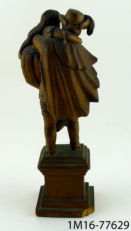 Träskulptur "Gustaf II Adolf bär moder Svea". Avbildar Gustaf II Adolf bärande moder Svea. Gustaf II Adolf står på en fyrkantig piedestal. Båda de avbildade personerna har på sig 1600talskläder.