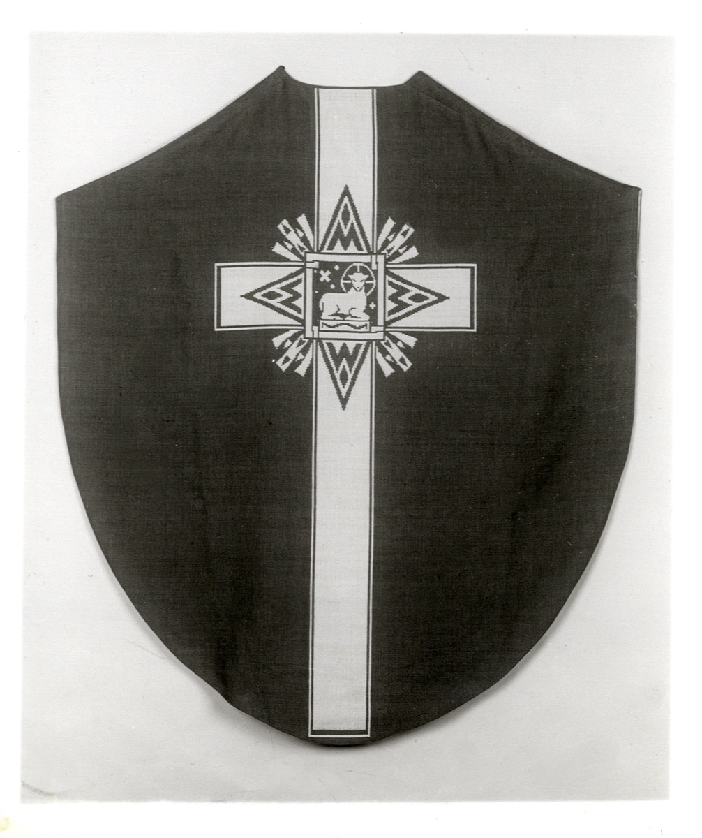 Foto (svart/vitt) av en mörk mässhake (framsidan) med ljusare broderat parti, med ett lamm med gloria. 

Inskrivet i huvudbok 1983.
