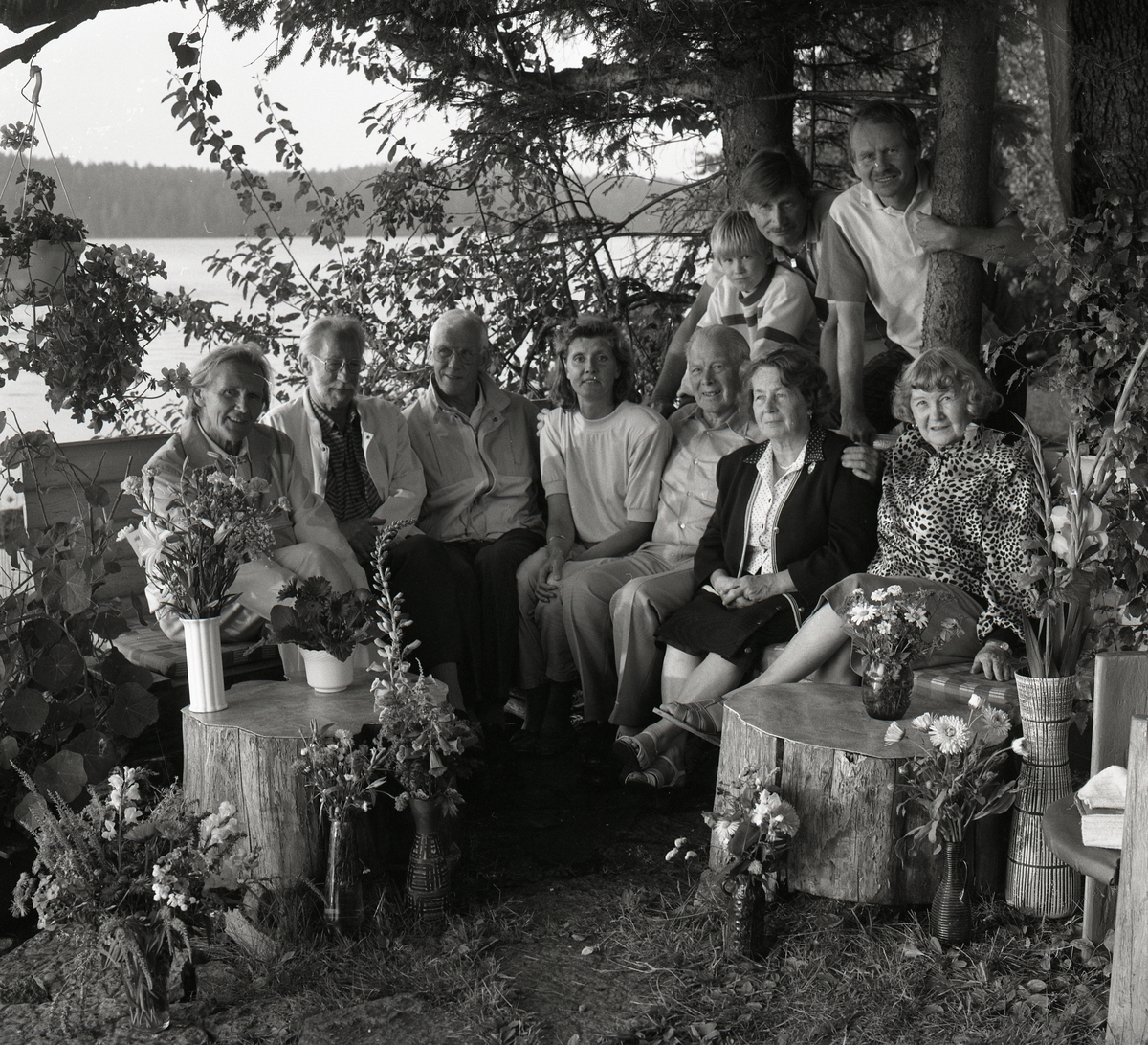 Hilding och Adéle sitter med några till människor under ett träd intill vattnet. Det hänger blommor i trädet och det är blommor i vaser som står på stubbarna. 80-årsdag 14 augusti 1990.