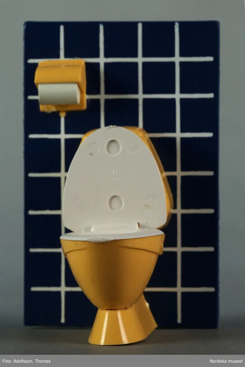 a) Toalettstol och toalettpappershållare, båda av gul och vit plast, som sitter fästade på en bit masonit, föreställande badrumsvägg av blått kakel med vita skarvar samt b) en badborste av vit och svart plast.