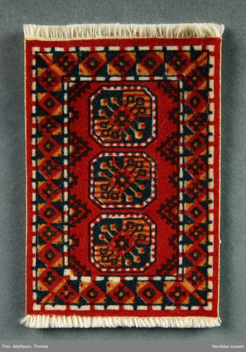 Wiltonmatta med orientaliskt tryckt mönster i röda nyanser. Hör till soffgruppen (NM.0333279 a-c) i hallen på första våningen i dockskåpet NM.0331721+.