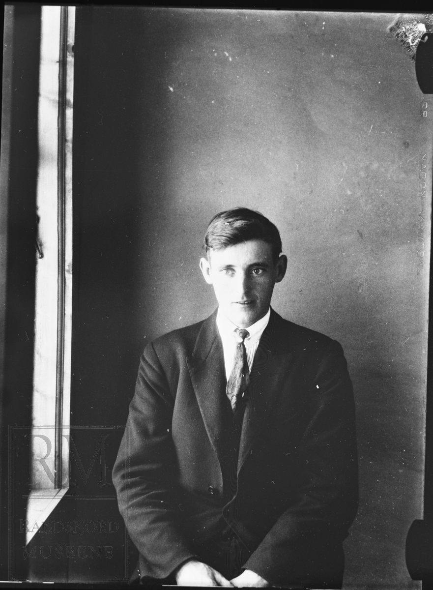 Portrett av ung mann sittende i dress, ved siden av et vindu.