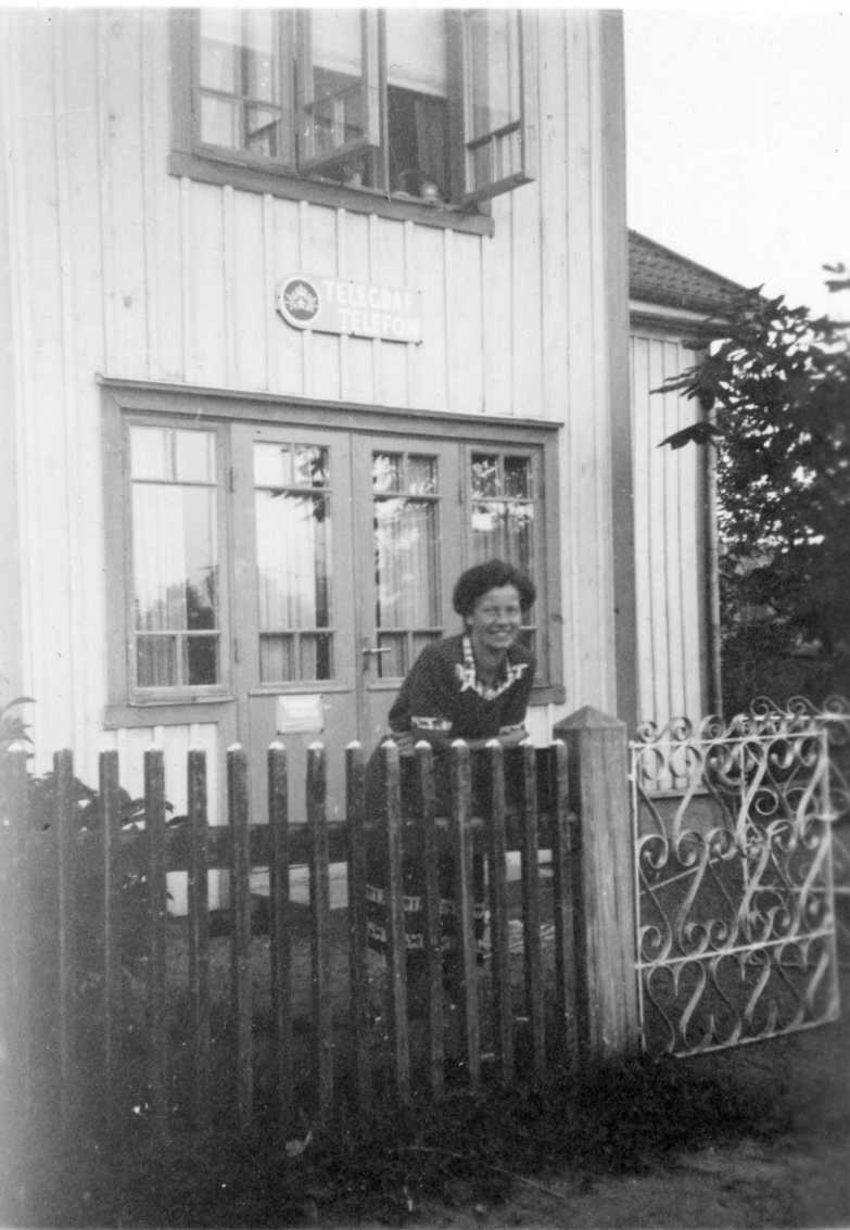 Birgit Englund står inne på en tomt lutad mot ett spjälstaket. På husväggen ovanför en entrédörr sitter en skylt: "Telegraf Telefon".
