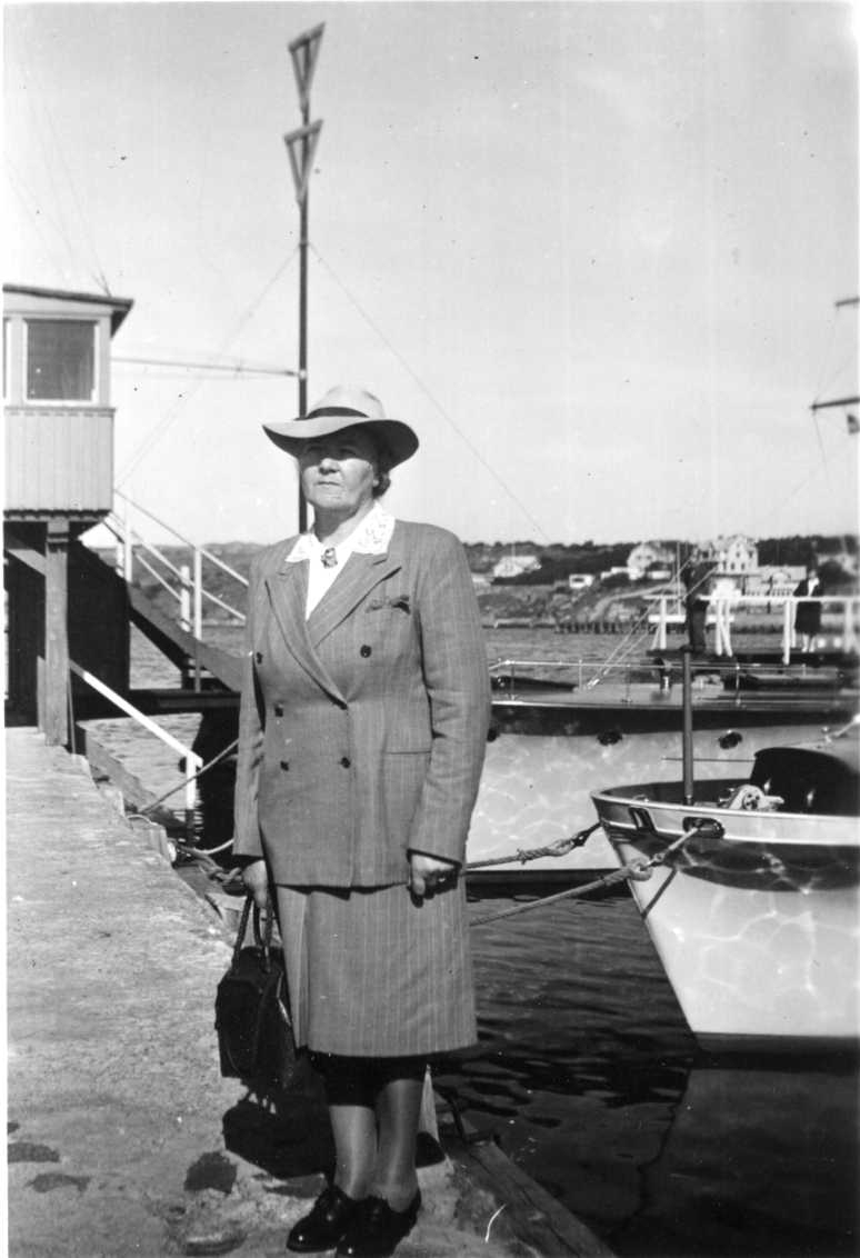 Ester Svensson med hatt kritsstrecksrandig dräkt, handväska i höger hand, står på en kaj. Båtar i bakgrunden.