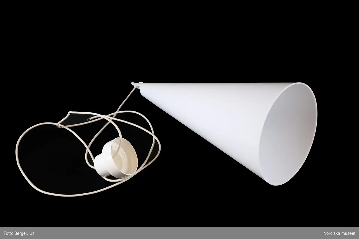 a) Taklampa av akrylplast, skärmen en långsmal vit kon. Vertikal skarv på den ena sidan markerad med en liten v-form vid mynningen. Upptill en mjukt böjd spets med två hål för sladden som gör att b) glödlampan kan höjas och sänkas efter behov. Vit plastsladd och takkopp.
Detta är den största modellen av Struten.
/Maria Maxén 2018-04-24