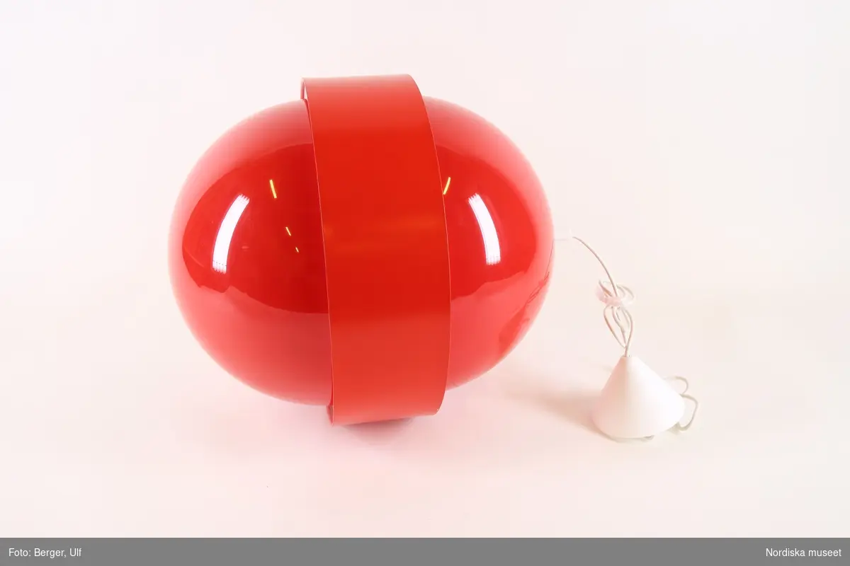 a-b) Taklampa av röd blank akrylplast, stående oval form med ett förkroppat band av vinyl runt mitten. Vit plastsladd, konisk takkopp av vit plast.
/Maria Maxén 2017-06-27