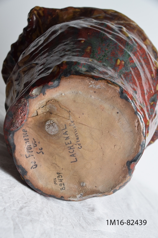 Oxblodsglaserad lergodskruka. Krukan är större upptill än i botten. Urnan visar relief med växtmotiv. Urnan har en skada upptill. Signatur i botten, signerad de Frumerie och Lachenal.
