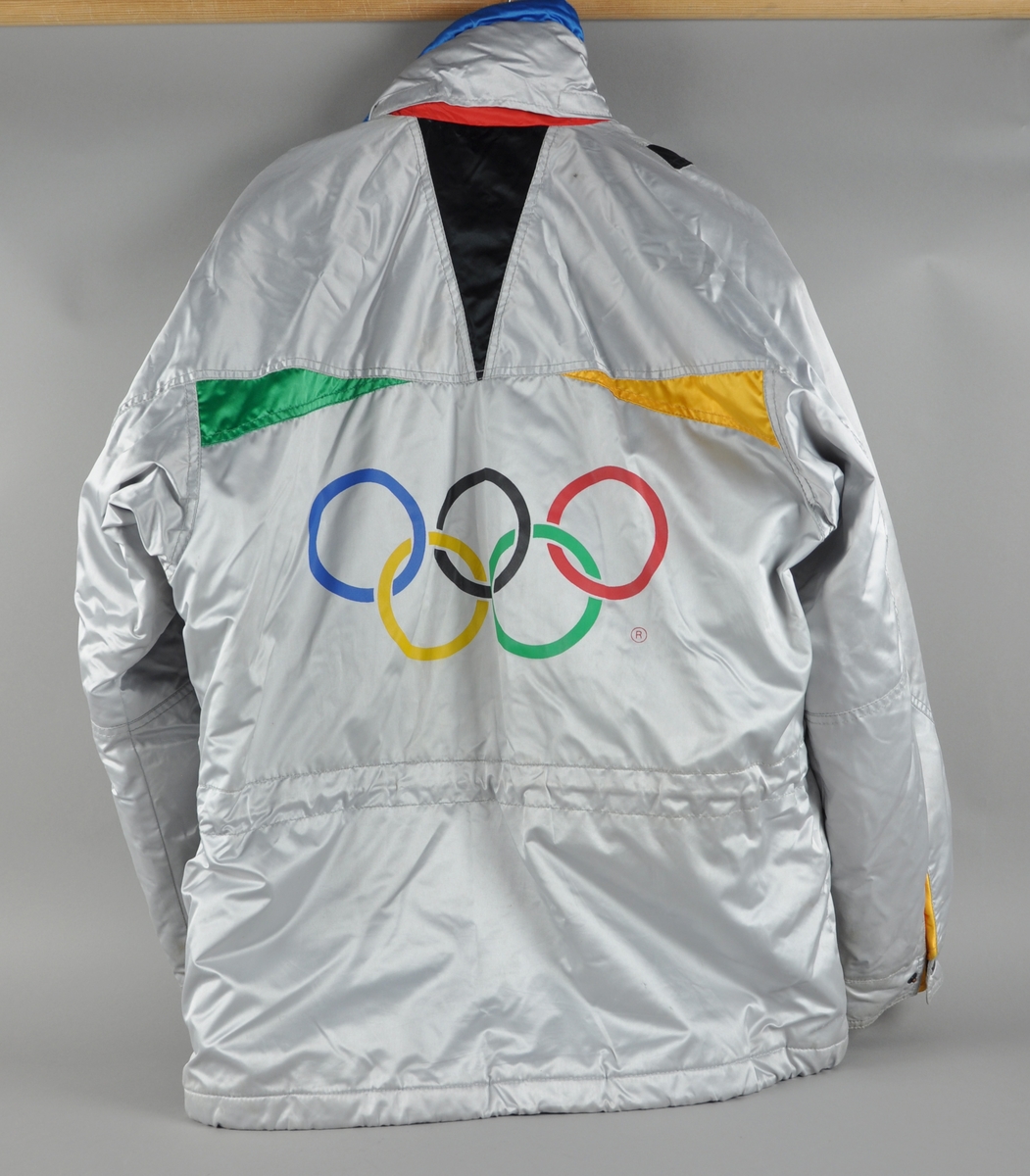 Grå jakke med dekor i ulike farger blant annet ved lommer og krage. Påsydd merker med logo for K-WAY og emblemet for de olympiske vinterleker i Albertville i 1992.