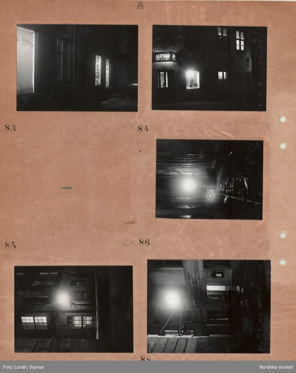 Motiv: (ingen anteckning) ;
Nattbild av stad med gator och byggnader möjligen Gamla Stan, husfasader med upplysta fönster, gatlyktor, skylt "Matvaruaffär",