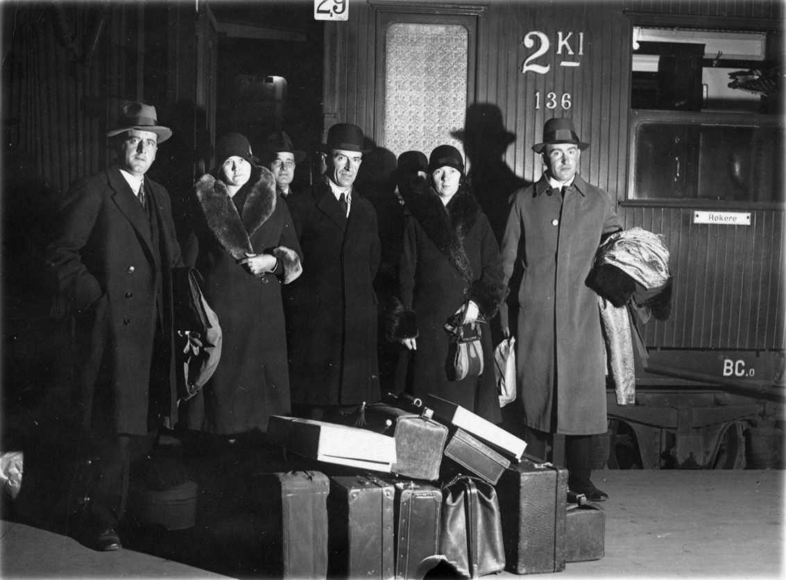 Ett sällskap herrar och damer i ytterkläder står vid en 2Kl-tågvagn. Framför dem på marken står det resväskor. Mannen i mitten är Gunnar Eliassen och till höger står Severin Skjelten.