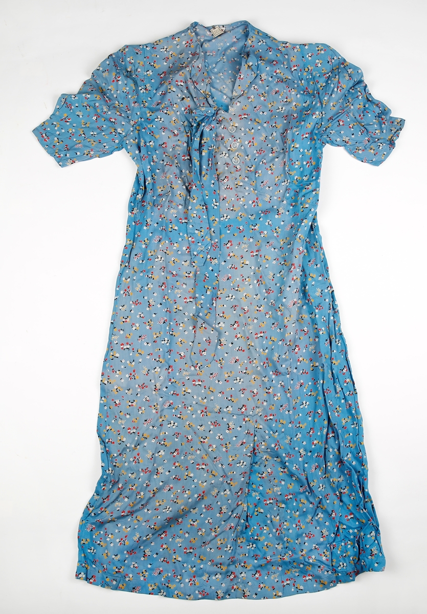 Blomstrete kjole i 1930-40-talssstil. Er sannsynligvis ubrukt da prislappen sitter i nakken. Korte ermer. Bånd i samme stoff følger med. Ser for kort ut til å være belte. Ermfisk i stoff på begge skuldre inni. Noe falmet i stoffet.