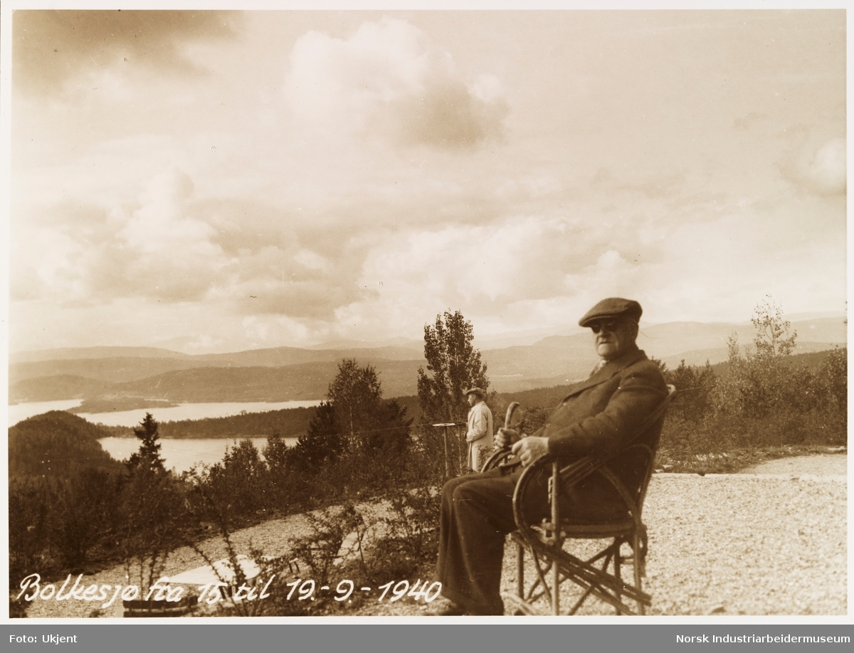 Mann med hatt, jakke og solbriller sitter i stol. En annen mann står i bakgrunnen. Utsikt ved Bolkesjø hotell