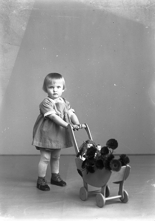 En liten flicka med dockvagn i en fotoateljé. I dockvagnen sitter en docka bland blommor. Flickan heter Ingrid Margareta Gustafsson boende i Gränna.