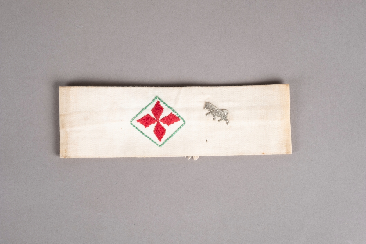 Et armbind i hvit bomull med rødt kors brodert i midten. Ved korset er det påsydd et merke av en hund.
