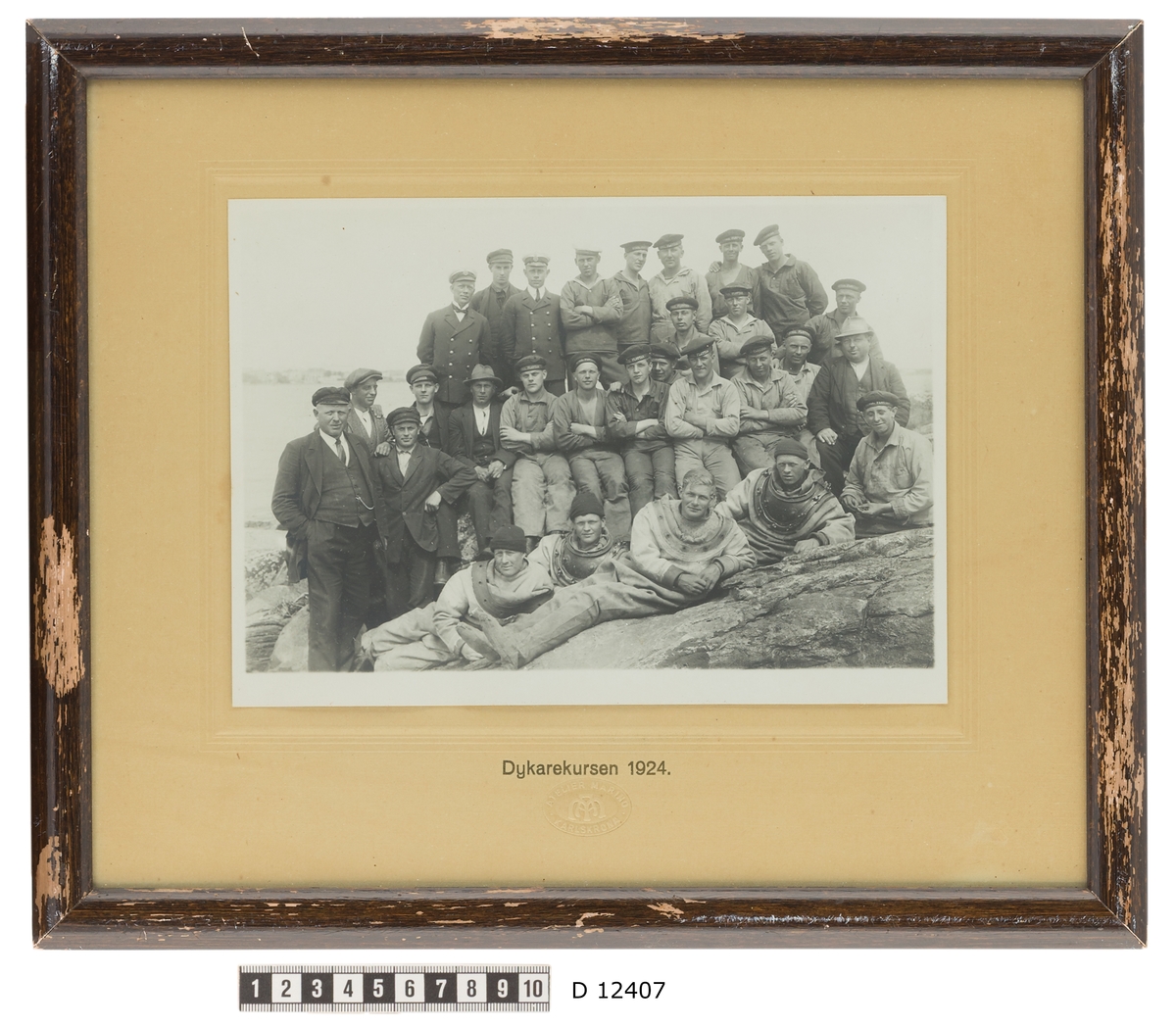 Denna grupporträtt föreställer dykarekursen 1924. En grupp marinkårssoldater har tagit plats på en klippa. I första raden ligger fyra män som bär tungdykardräkt. I andra raden är det arbetskläder som gäller och högst upp bär man sjömanskostym.