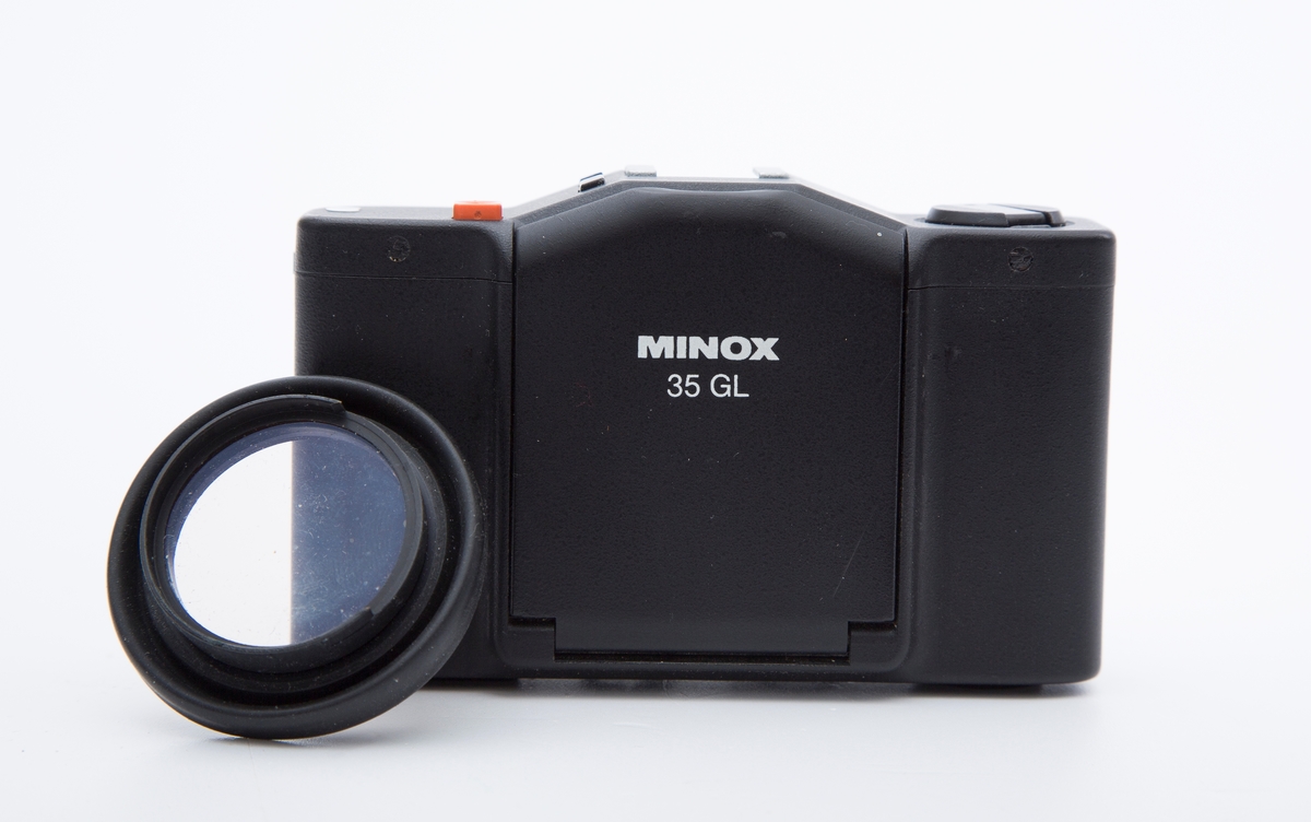 Kamera i sort skinnetui og beskyttelseslinse i skinnetui med rem satt fast på kamera. Rektangulært kamera. Mrk:"Minox 35 GL". 