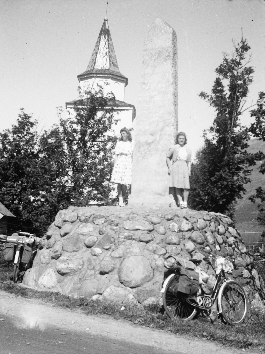 Motiv av to kvinner som står ved et monument framfor ei kirke.