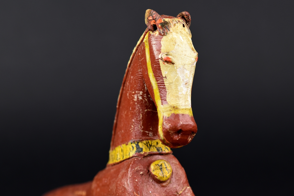 Käpphäst av trä. 
Hästen är målad i brunt och vitt, seldonen är målade i gult. 
Huvud, manke och framben utgör den främre delen av käpphästen. 
Öronen är tillverkade av läder.